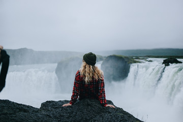 Top view on young female with blonde hair under green beanie hat and in red checkered shirt sit on edge of epic giant waterfall in iceland during midnight summer sun. Amazing adventure of lifetime