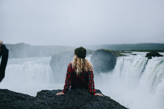 Top view on young female with blonde hair under green beanie hat and in red checkered shirt sit on edge of epic giant waterfall in iceland during midnight summer sun. Amazing adventure of lifetime