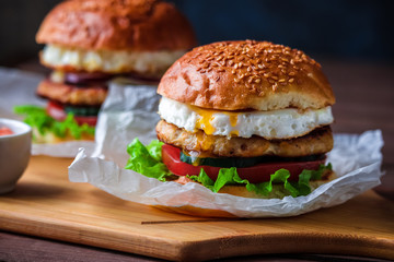 homemade burgers with chicken cutlet fried egg dripping yolk tomato lettuce cucumber on cutting board close up
