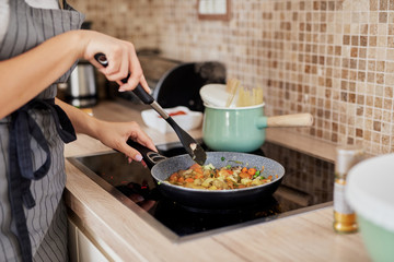 Charming Caucasain brunette in apron standing next to stove in kitchen and cooking healthy dinner.