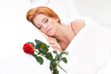 Beautiful young woman is sleeping on white bed. Flower rose on pillow next to her. Closeup portrait of attractive Girl with red hair, closed eyes - indoors.