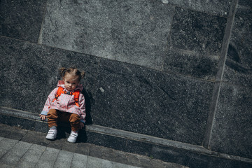 little pretty baby who got lost in the city, a girl in a pink raincoat, with a backpack and a stylish hairstyle sitting next to a large administrative building one by one