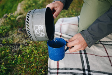 Hands of male adventurer tourist or hiker at camping site pour boiling water from fast burner or outdoor gas stove pot into metal mug or cup to make hot beverage coffee or tea for breakfast in tent