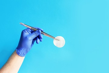 Medicine and Surgery theme: doctor's hand in a blue glove holding a pair of tweezers with a cotton pad on a blue background, isolated. - obrazy, fototapety, plakaty