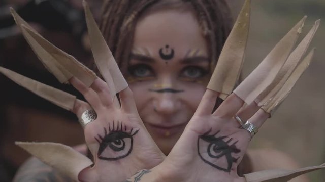 Hands of dryad or forest fairy with painted eyes on the palms and false claws dancing and covering face. Ancient ritual of forest creatures. Performance of dancers in forest