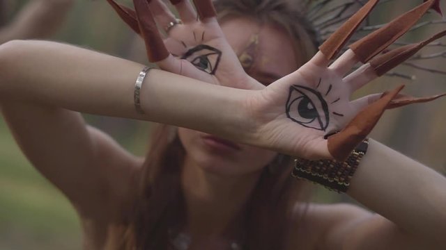 Hands of dryad or forest fairy with painted eyes on the palms and false claws dancing and covering face. Ancient ritual of forest creatures. Performance of dancers in forest