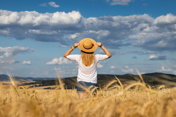 Happy woman with straw hat standing in wheat field and enjoying summer. Blond hair woman feeling freedom. 