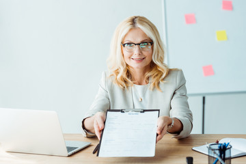 blonde cheerful woman in glasses holding clipboard with resume while sitting near desk