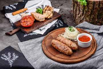 Fried meat sausages, served with pickled onions and barbecue sauce on a round wooden board on dark concrete background