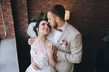 Stylish groom gently hugs a beautiful brunette bride against the background of a brick wall. Wedding portrait of newlyweds in love in a modern studio with a beautiful interior. Conceptual photography.