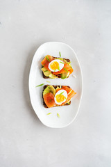 Healthy open sandwiches on multigrain wholegrain  toast with avocado, salmon, eggs, herbs, sunflower seeds over white plate on concrete background with copy space. Healthy protein food.