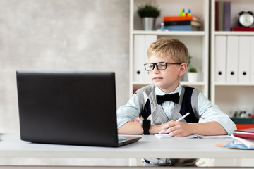 Blond boy in casual clothing and glasses sits behind desk and looking at laptop