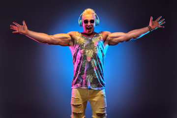 Obraz premium Fashion Muscular DJ man listen to music in colorful neon light with hands up. Handsome blonde Hipster guy Having Fun happy. Art neon fashionable style. Music festival nightclub, summer party concept