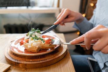 Traditional Belgian waffle with salmon, lettuce leaves and poached egg. A young woman is having Breakfast in a cafe, hands with a fork and a knife close-up.