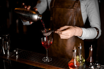 Bartender in leather apron pouring a red alcoholic drink from the steel shaker through the sieve