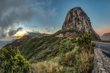 Amazing sunrise near Los roques and famous Agando rock near Garajonay Park at La Gomera. Half moon on a background of gray and purple clouds. Relic laurels and heather on steep slopes. Canary, Spain