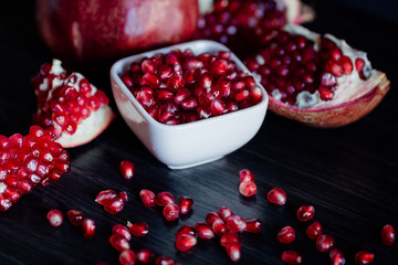 White bowl with pomegranate seeds and ripe pomegranate on dark wooden background.