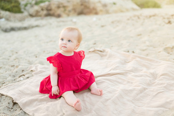 Cute baby girl in a red dress sitting and playing on the beach with sand