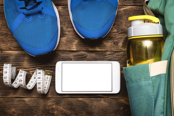 Mobile phone with a blank screen, blue sneakers and a backpack with a bottle of water on a wooden board background.
