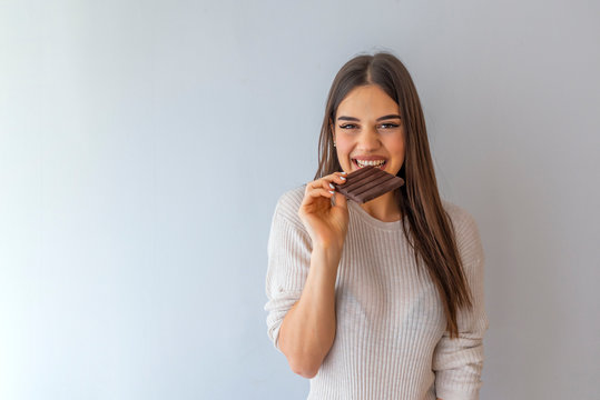  Young woman with natural make up having fun and eating chocolate isolated on gray background. Woman eating chocolate. Happy teenage girl eating chocolate bar