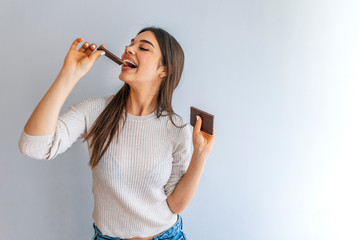 Portrait of a happy young woman eating chocolate bar isolated over grey background. Happy young beautiful lady eating chocolate and smiling. Girl tasting sweet chocolate.