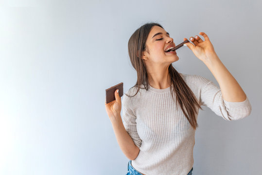 Portrait of a happy young woman biting chocolate bar isolated over grey background. Health, people, food and beauty concept - Lovely smiling teenage girl eating chocolate