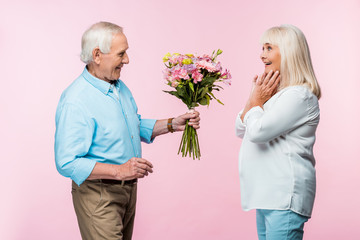 cheerful senior man giving bouquet with flowers to excited wife on pink