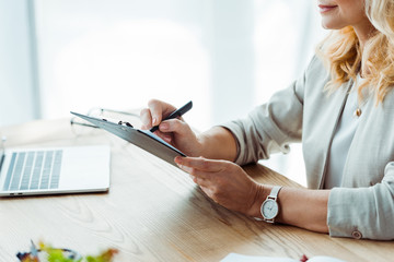 cropped view of woman writing while holding clipboard in office