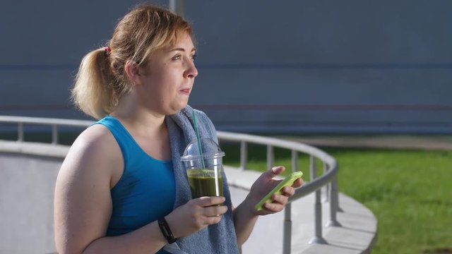 Close-up of body positive slimming female jogger drinking healthy green smoothie and using cellphone after workout. Plus size woman losing weight, sport lifestyle, healthy balanced diet and drinks