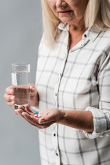 cropped view of senior woman holding pills and glass of water isolated on grey