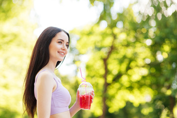 Young beautiful girl drinking fresh juice from plastic cup take-out food. Smiling slim brunette woman with berry lemonade in park. Summer cold drinks.