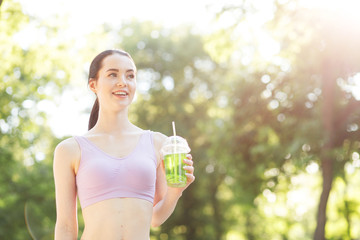 Young beautiful girl drinking green beverage with lime and kiwi from plastic cup take-out food. Smiling slim brunette woman with lemonade in park. Summer cold drinks.