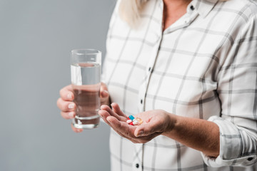 cropped view of retired woman holding pills and glass of water isolated on grey