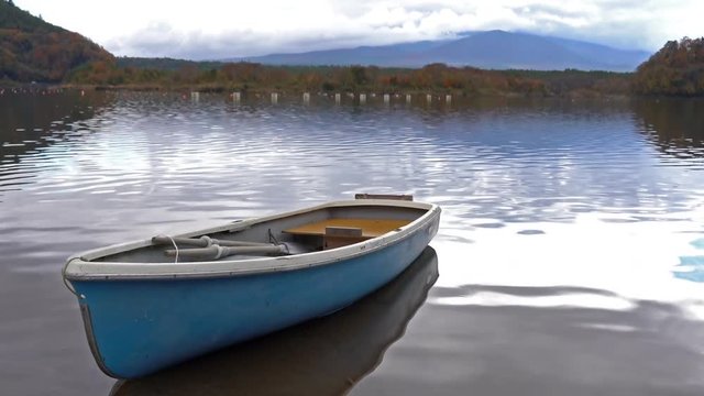 A small blue and white rowboat sits moored in the sandy bottom of a quiet lake surrounded​ by tall mountains and fall colored trees
