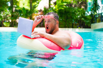 surprised Fat funny man in pink inflatable circle in pink glasses works on a laptop in a swimming pool