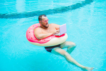 surprised Fat funny man in pink inflatable circle in pink glasses works on a laptop in a swimming pool