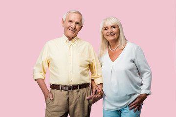 happy retired man with hand in pocket holding hands with cheerful wife isolated on pink