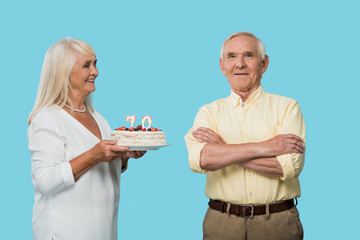 cheerful retired woman holding birthday cake near happy husband with crossed arms isolated on blue