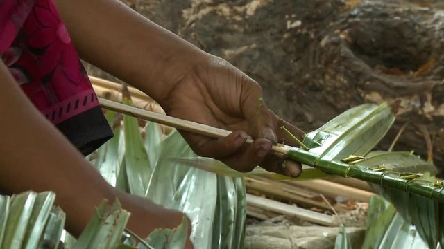 Extreme close-up low-angle tilting shot of a woman preparing a nipa palm roof thatch. Here hands are folding, bending, and hooking the fresh green leaves systematically on a smooth stick, Rakhine