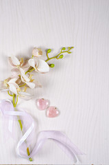 Plastic orchid sprig with the greenish stem wrapped by silk ribbons and two rose quartz hearts lay on a light gray wooden background. Top view, copy space.