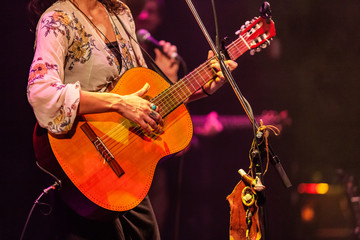 Band member uses guitar and mic stand. A close up view on a stylish lady band member playing a guitar during a live music set. Blurred musicians are seen in the background. Details on stage at gig.