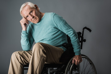 Fototapeta na wymiar upset retired man sitting in wheelchair and looking at camera on grey