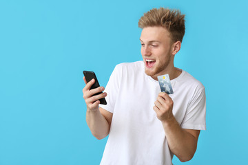 Excited young man with credit card and mobile phone on color background