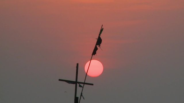Close-up low-angle sunset still shot of a beautiful ball of orange sun above a silhouette of a boat's cross and flag-post, Ngapali beach, Myanmar