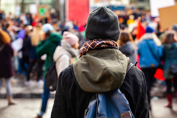 Person watches ecological rally. A young make is viewed from the back, wearing a hat and backpack,...