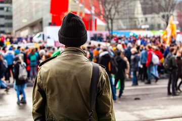 Man watches people march for environment. A young man is seen from the back, wearing an olive jacket and wooly hat, as eco-activists march against global warming in a blurry background.
