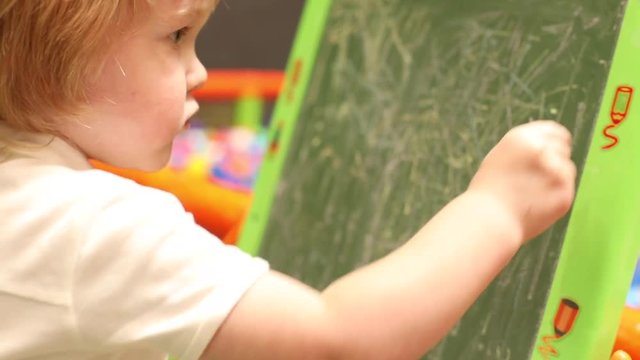 Closeup creative caucasian male child drawing with chalk on blackboard in modern children entertainment center. Cute boy holding colored chalks. Concentrated face slow motion view. Young talent artist