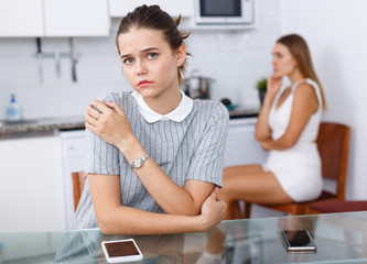 Sad girl sitting at table after conflict, woman friend on backround
