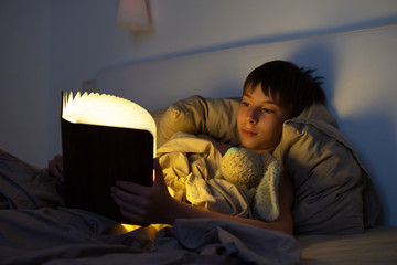 Adorable boy reads book before sleeping in bed at home with his old toy. Happy childhood.