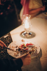 Stylish happy bride and groom tasting together wedding cake with fruits at wedding reception outdoors in the evening. Couple  eating cake in soft candle light in night.